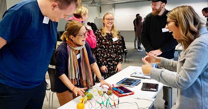 Beim Netzwerktreffen von KuMuS am 18. Oktober 2024 testen Teilnehmer an einem Tisch ein interaktives Setup mit Früchten, die als Sensoren fungieren, verbunden mit Tablets und Kabeln. Eine Frau erklärt begeistert die Funktionsweise, während andere lächeln und interessiert zuhören. Im Hintergrund sind weitere Besucher in Gespräche vertieft und erkunden beim offenen Austausschformat den Markt der Möglichkeiten.